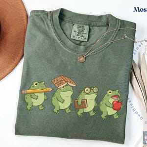 K&ouml;nnte beinhalten: Ein moosgr&uuml;nes T-Shirt mit einer Grafik von vier Zeichentrickfr&ouml;schen. Ein Frosch h&auml;lt einen gelben Bleistift, einer hat ein Buch mit dem Titel Nature Study auf dem Kopf, einer tr&auml;gt eine Brille und ein Buch, und einer h&auml;lt einen roten Apfel.