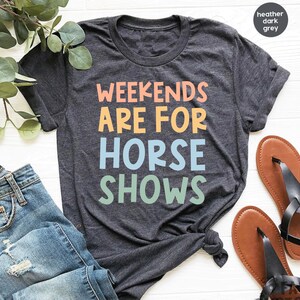 May include: Dark gray t-shirt with the text "Weekends Are For Horse Shows" in colorful letters. The shirt is paired with blue jeans and brown sandals, suggesting a casual, equestrian-themed style.
