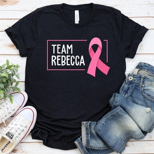 Pode incluir: Uma camiseta preta com o texto "TEAM REBECCA" em uma caixa branca e uma fita rosa. A camiseta est&aacute; colocada sobre uma superf&iacute;cie de madeira branca com um par de t&ecirc;nis brancos e uma cal&ccedil;a jeans azul.