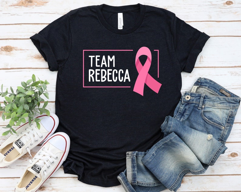 Pode incluir: Uma camiseta preta com o texto "TEAM REBECCA" em uma caixa branca e uma fita rosa. A camiseta est&aacute; colocada sobre uma superf&iacute;cie de madeira branca com um par de t&ecirc;nis brancos e uma cal&ccedil;a jeans azul.