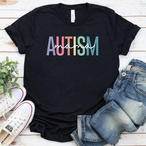 May include: A black t-shirt with the text "AUTISM mama" in rainbow colors. The shirt is laid flat on a white wooden surface with a pair of white sneakers and blue jeans.