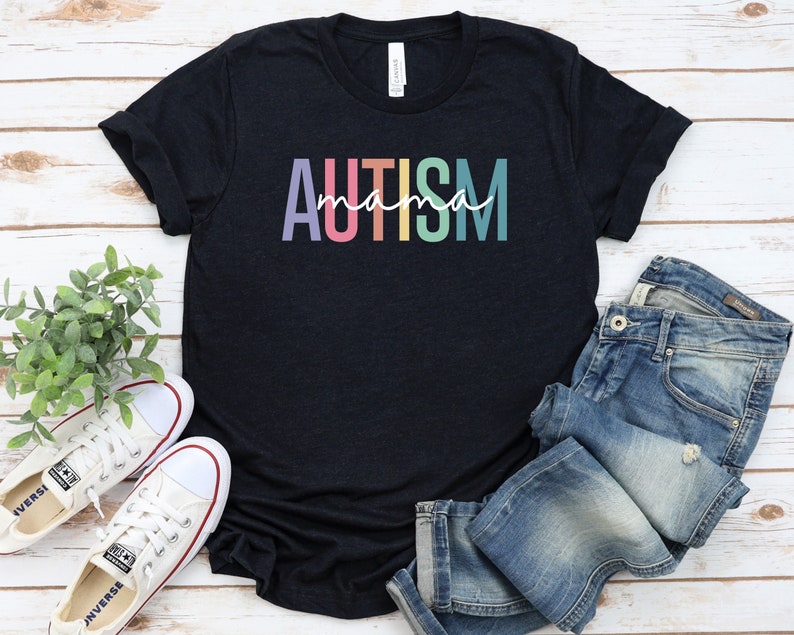 May include: A black t-shirt with the text "AUTISM mama" in rainbow colors. The shirt is laid flat on a white wooden surface with a pair of white sneakers and blue jeans.