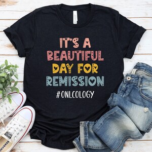 May include: Black t-shirt with the text "It's a beautiful day for remission #oncology" in colorful letters with hearts. The shirt is on a wooden surface with a pair of white sneakers and blue jeans.