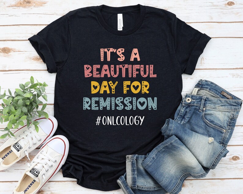 May include: Black t-shirt with the text "It's a beautiful day for remission #oncology" in colorful letters with hearts. The shirt is on a wooden surface with a pair of white sneakers and blue jeans.