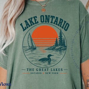 May include: A moss green t-shirt featuring a circular graphic of Lake Ontario. The design includes a sailboat, a loon, pine trees, and a large orange sun. Text reads LAKE ONTARIO, THE GREAT LAKES, ONTARIO, and NEW YORK in dark blue and orange.