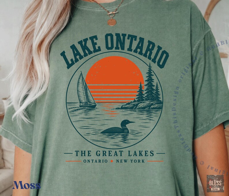 May include: A moss green t-shirt featuring a circular graphic of Lake Ontario. The design includes a sailboat, a loon, pine trees, and a large orange sun. Text reads LAKE ONTARIO, THE GREAT LAKES, ONTARIO, and NEW YORK in dark blue and orange.