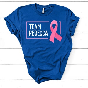 Pode incluir: Camiseta azul com o texto "Team Rebecca" em uma caixa branca e uma fita rosa.