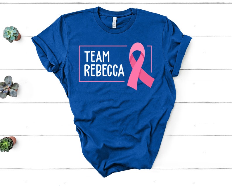 Pode incluir: Camiseta azul com o texto "Team Rebecca" em uma caixa branca e uma fita rosa.
