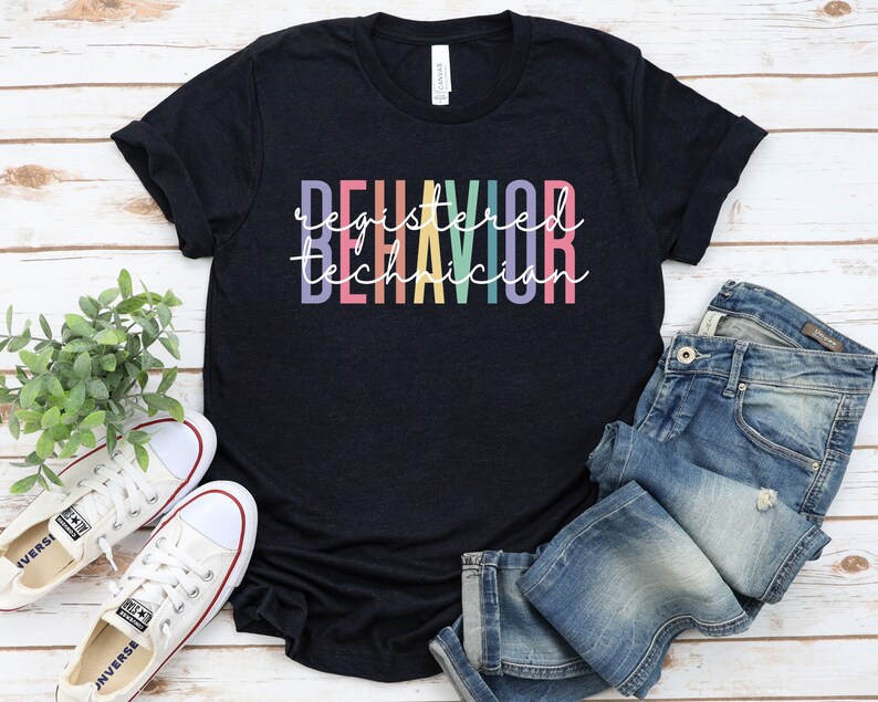 Puede incluir: Una camiseta negra con el texto "Registered Behavior Technician" en colores arco&iacute;ris.