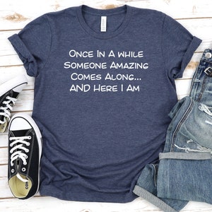 Peut inclure: Un t-shirt bleu marine avec un texte blanc qui dit "ONCE IN A WHILE SOMEONE AMAZING COMES ALONG... AND HERE I AM".