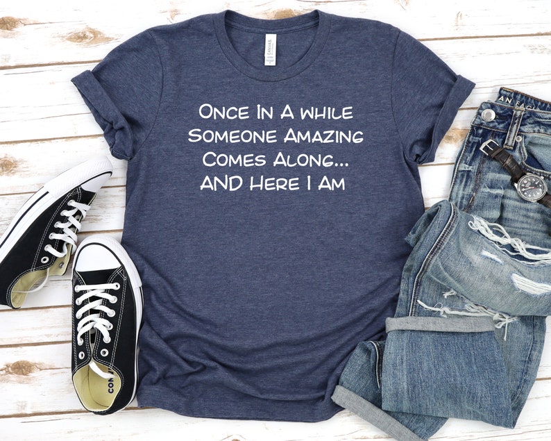 Peut inclure: Un t-shirt bleu marine avec un texte blanc qui dit "ONCE IN A WHILE SOMEONE AMAZING COMES ALONG... AND HERE I AM".