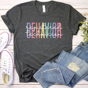 Puede incluir: Una camiseta gris oscuro con el texto "registered behavior technician" en colores arco&iacute;ris.