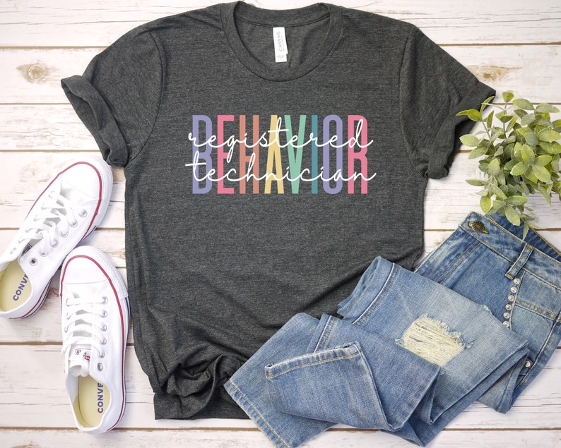 Puede incluir: Una camiseta gris oscuro con el texto "registered behavior technician" en colores arco&iacute;ris.