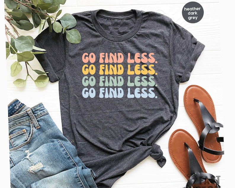 May include: A heather dark grey t-shirt with the text "GO FIND LESS." repeated four times in a rainbow color scheme. The text is in a retro font style.