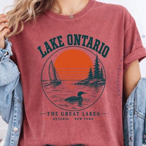 May include: A brick-red short-sleeve t-shirt featuring a circular graphic with a sailboat, a loon, pine trees, and an orange sun. The text reads LAKE ONTARIO, THE GREAT LAKES, and ONTARIO NEW YORK. The shirt has a crew neckline and a vintage-style finish.