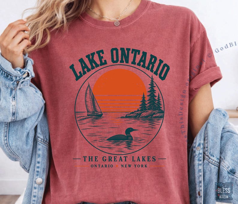 May include: A brick-red short-sleeve t-shirt featuring a circular graphic with a sailboat, a loon, pine trees, and an orange sun. The text reads LAKE ONTARIO, THE GREAT LAKES, and ONTARIO NEW YORK. The shirt has a crew neckline and a vintage-style finish.