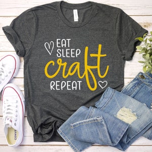 May include: A dark gray t-shirt with the text "Eat Sleep Craft Repeat" in white and yellow lettering. The text is surrounded by two small white hearts.