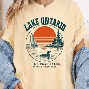 May include: A pale yellow t-shirt featuring a circular graphic with a sailboat, a loon on the water, and pine trees against a large orange sun. The text reads LAKE ONTARIO, THE GREAT LAKES, and ONTARIO NEW YORK in dark green and orange lettering.
