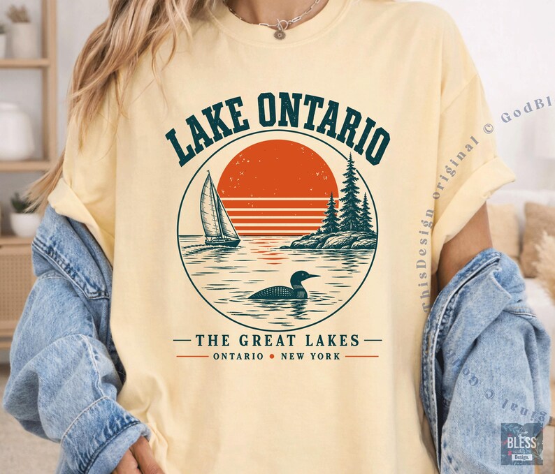 May include: A pale yellow t-shirt featuring a circular graphic with a sailboat, a loon on the water, and pine trees against a large orange sun. The text reads LAKE ONTARIO, THE GREAT LAKES, and ONTARIO NEW YORK in dark green and orange lettering.