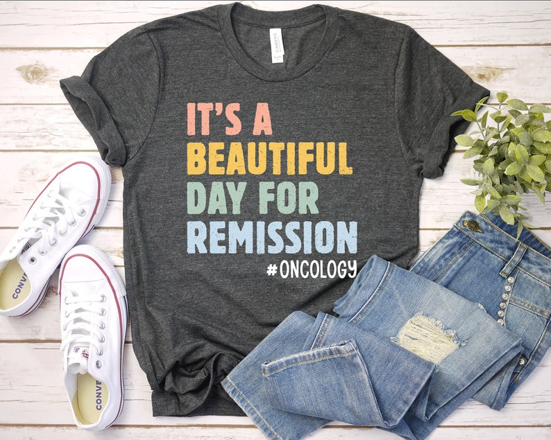 May include: A dark gray t-shirt with the text "It's a beautiful day for remission #oncology" in rainbow colors. The shirt is laid out on a white wooden surface with a pair of white sneakers and blue jeans.
