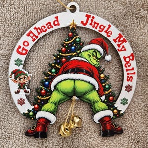 May include: A circular white wooden Christmas ornament featuring a green character in a Santa suit bending over in front of a decorated tree. The text Go Ahead Jingle My Bells is printed in red. A small elf figure and two gold metal bells are attached.