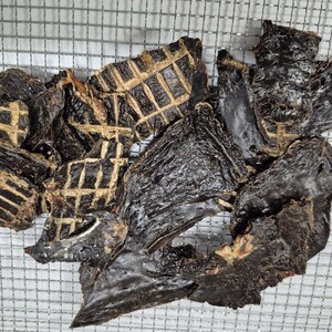 May include: A pile of dark brown, dried beef jerky pieces. Some pieces have a grid pattern on them.