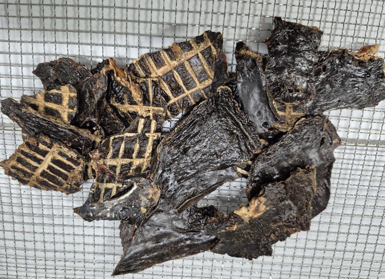 May include: A pile of dark brown, dried beef jerky pieces. Some pieces have a grid pattern on them.