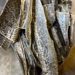 Puede incluir: Primer plano de trozos de piel de pescado seca. Las piezas var&iacute;an en tama&ntilde;o y forma, con una mezcla de tonos marrones claros y oscuros. La textura parece escamosa y ligeramente curvada, lo que sugiere una calidad crujiente o quebradiza.