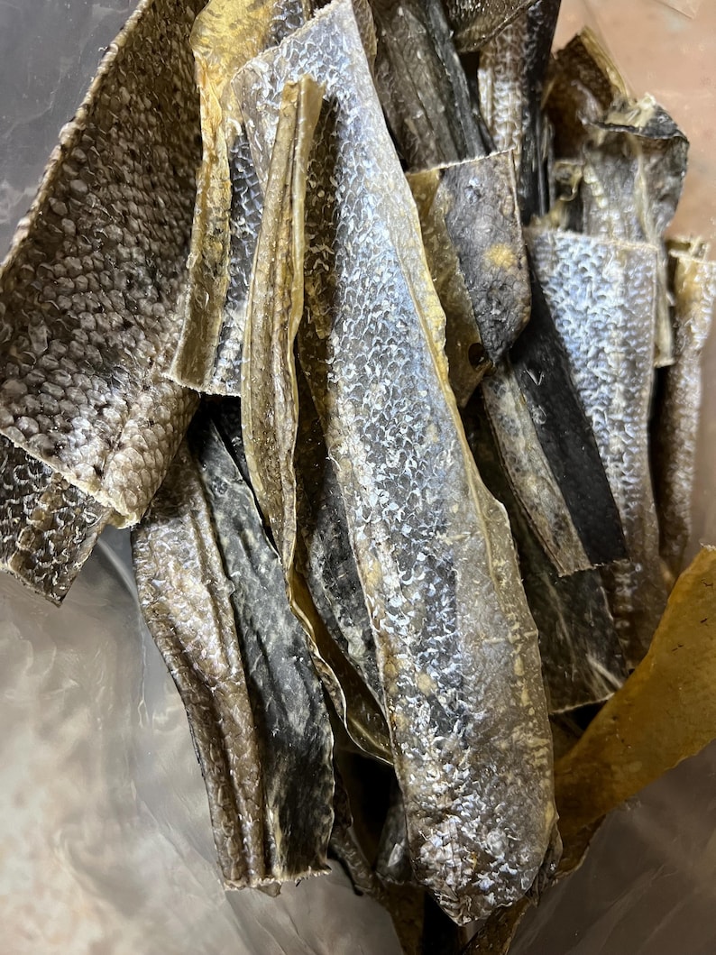 Puede incluir: Primer plano de trozos de piel de pescado seca. Las piezas var&iacute;an en tama&ntilde;o y forma, con una mezcla de tonos marrones claros y oscuros. La textura parece escamosa y ligeramente curvada, lo que sugiere una calidad crujiente o quebradiza.