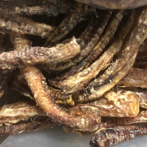 Può includere: Primo piano di un mucchio di snack per cani secchi, marroni e marroncini. Gli snack hanno una forma tubolare con una superficie strutturata e sono fittamente impacchettati. Gli snack sembrano essere un prodotto naturale.