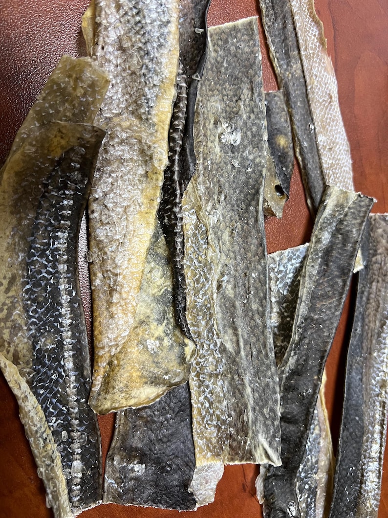 Puede incluir: Primer plano de trozos de piel de pescado seca. Las piezas var&iacute;an en tama&ntilde;o y forma, con una mezcla de colores beige claro, gris y negro. La textura parece &aacute;spera y escamosa, con algunas piezas que muestran la espina.