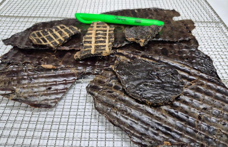 May include: A close-up of a pile of dark brown, dried meat treats on a wire rack. The treats are in various shapes and sizes, some are flat and others are thicker and more textured. A green highlighter pen is lying on top of the pile.