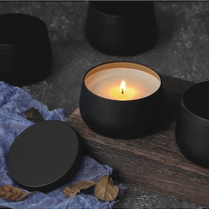 Puede incluir: Una vela negra encendida en un recipiente redondo con interior dorado. La vela está sobre una superficie de madera con un paño azul y otros recipientes negros en el fondo. La llama es brillante y la cera es blanca.