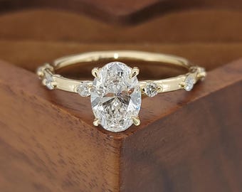 May include: A gold engagement ring with an oval-cut diamond center stone. The band features small accent stones and a delicate design. The ring is set against a wooden background, showcasing its elegant details.