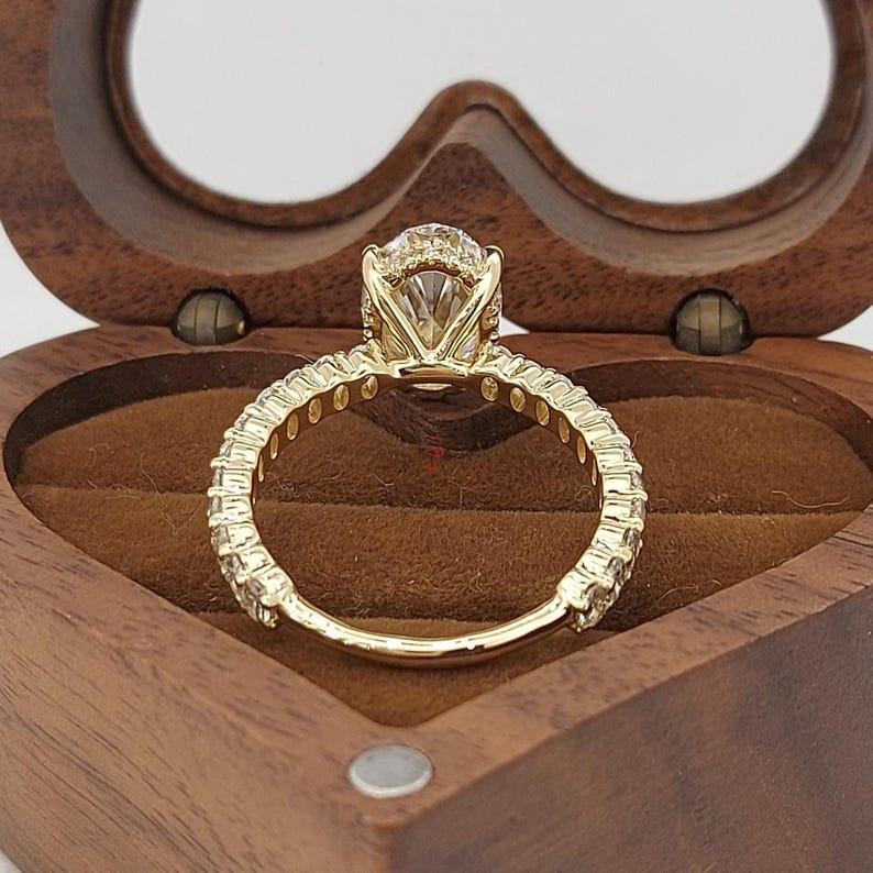 May include: A gold engagement ring with a large, clear center stone and smaller stones set along the band. The ring is displayed in a heart-shaped wooden box with a brown interior.