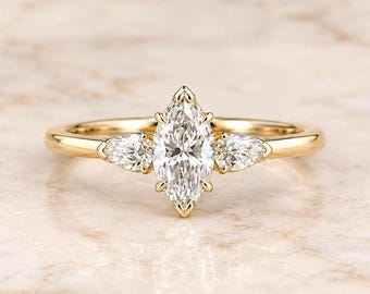 May include: A gold ring featuring a central marquise-cut clear gemstone flanked by two smaller pear-cut clear gemstones. The band is smooth and polished, holding the three stones in a prong setting that highlights the clarity of the gems.