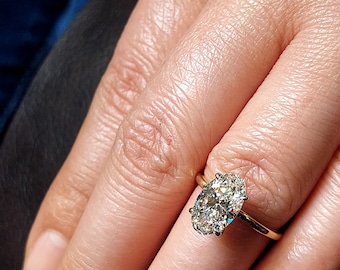 May include: A gold ring with an oval-shaped diamond. The diamond is set in a prong setting and has a brilliant cut. The ring is on a finger.