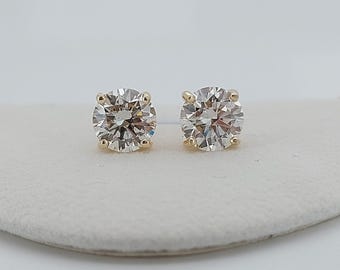 May include: A pair of round-cut diamond stud earrings. The diamonds are set in a four-prong, gold-colored setting. The earrings are displayed on a white surface against a white background. The diamonds have a brilliant cut.