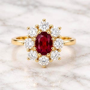 Op de afbeelding: Een geelgouden ring met een centrale ovaal geslepen dieprode robijn, omringd door een halo van tien ronde, briljant geslepen witte diamanten. De stenen zijn gezet in een bloemvormig cluster op een gepolijste gouden ring met een gladde afwerking.