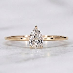 May include: A gold ring with a pear-cut diamond. The diamond is set on a slender, polished gold band. The ring is presented on a white surface, accentuating its brilliance. The band is approximately 1 mm thick.