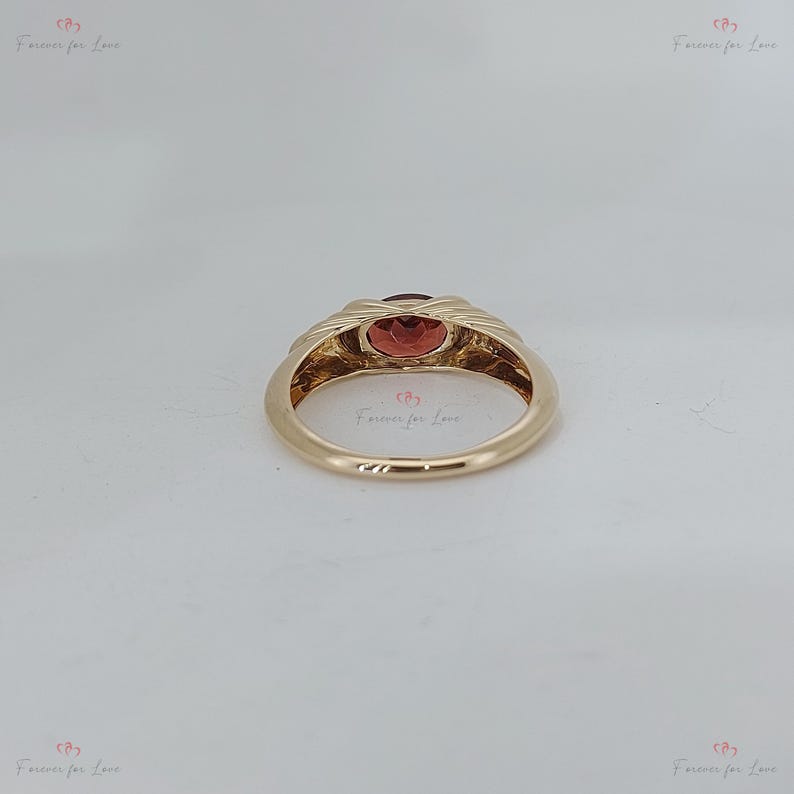 Puede incluir: Un anillo de oro con una piedra preciosa ovalada de color rojo anaranjado. La banda del anillo tiene un dise&ntilde;o texturizado y tejido alrededor de la piedra preciosa. El anillo est&aacute; sobre un fondo neutro. Las palabras "Forever for Love" est&aacute;n impresas en la imagen.