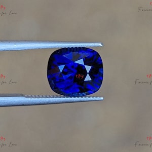 May include: A deep blue gemstone with a cushion cut, held by tweezers. The gemstone is faceted and has a brilliant shine.