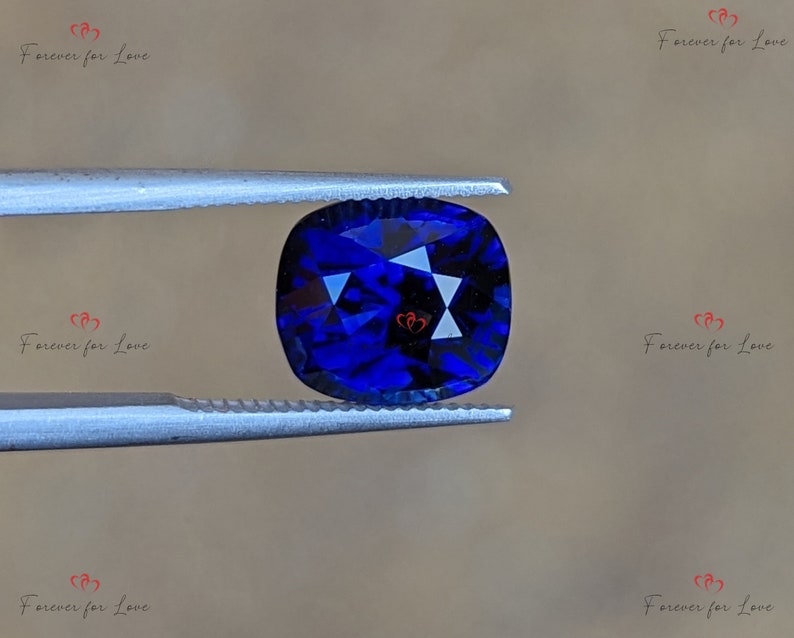 May include: A deep blue gemstone with a cushion cut, held by tweezers. The gemstone is faceted and has a brilliant shine.