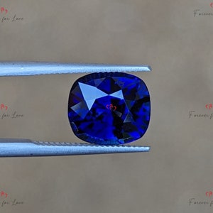 May include: A deep blue cushion-cut gemstone held by tweezers. The gemstone has a faceted cut and is a vibrant blue color.