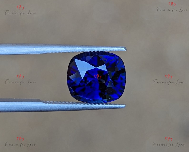 May include: A deep blue cushion-cut gemstone held by tweezers. The gemstone has a faceted cut and is a vibrant blue color.