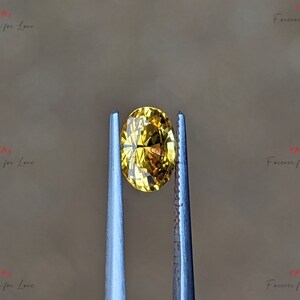 May include: A yellow oval-shaped gemstone held by tweezers. The gemstone is transparent and has a slight golden hue.