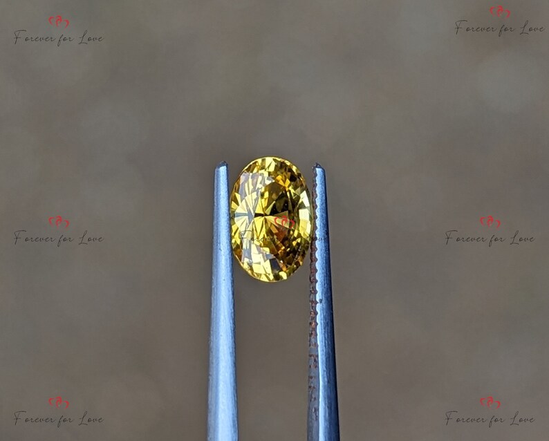 May include: A yellow oval-shaped gemstone held by tweezers. The gemstone is transparent and has a slight golden hue.