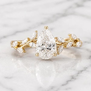 May include: A gold ring featuring a large, pear-shaped diamond as the centerpiece. The band is adorned with smaller, marquise-cut diamonds and a delicate leaf design, creating an elegant and nature-inspired aesthetic.