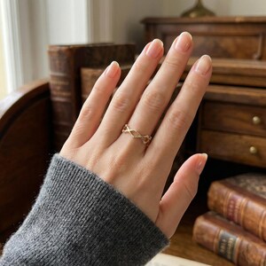May include: A gold ring with an open, interwoven design is shown on a hand. The ring's intricate pattern creates a delicate, geometric aesthetic. The hand is wearing a grey sweater, and the background includes antique books and wooden furniture.