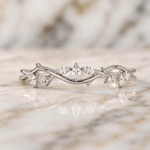 Könnte beinhalten: Ein silberfarbener Ring mit einer Ringschiene, die wie ineinander verschlungene Zweige gestaltet ist. Der Ring ist mit sieben kleinen, klaren, marquise-geschliffenen Steinen besetzt, die oben angeordnet sind und Blätter an einer Ranke imitieren.